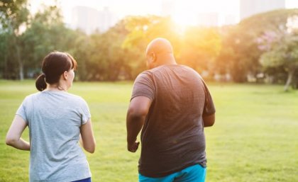 Two people, a man and woman, wear exercise gear outdoors in a green park and walk away from the camera.